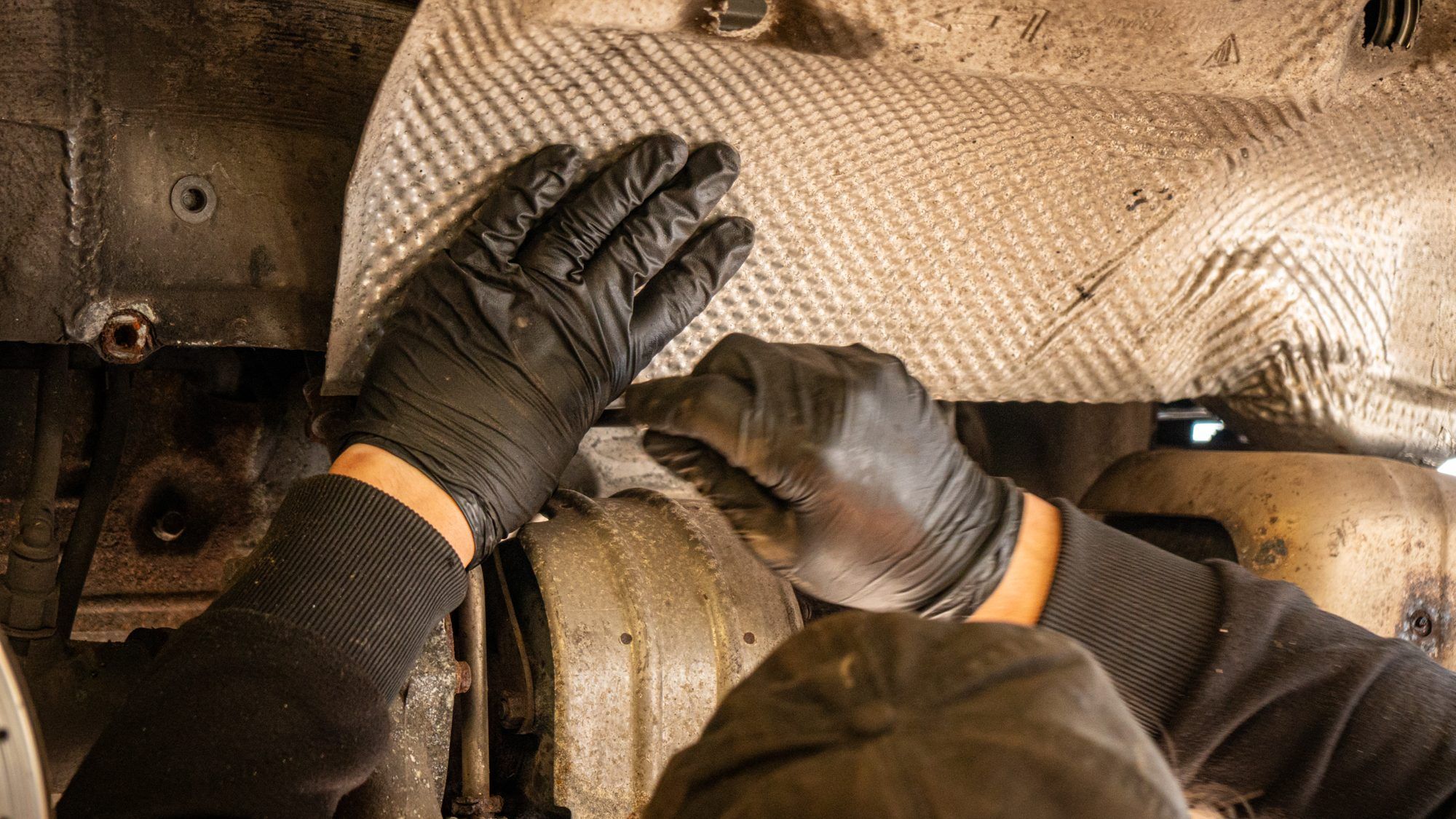Gloved hands fitting a heat shield under a Porsche